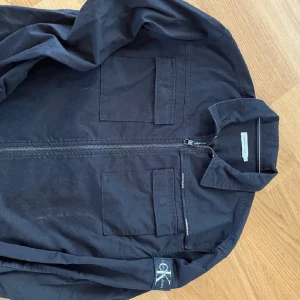 Svart jacka från Calvin Klein Jeans - Säljer en snygg svart jacka från Calvin Klein Jeans i storlek M. Jackan har en dragkedja framtill och två bröstfickor med lock. Perfekt för höst och vår, med en stilren design som passar till det mesta. Skicka ett meddelande vid intresse!