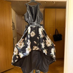 Svart blommig klänning - Elegant svart klänning med blommönster i vitt och beige. Klänningen har en hög-låg design med en ärmlös topp och en vid kjol. Perfekt för speciella tillfällen. Klänningen är 38 men kan även användas av personer med storlek 40