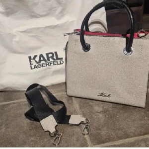 Handväska från Karl Lagerfeld - Elegant handväska från Karl Lagerfeld med svarta handtag och avtagbar axelrem. Väskan har en stilren design med logotyp framtill och dragkedja upptill. Perfekt för att ge en touch av lyx till din outfit. Dustbag medföljer vid köp.