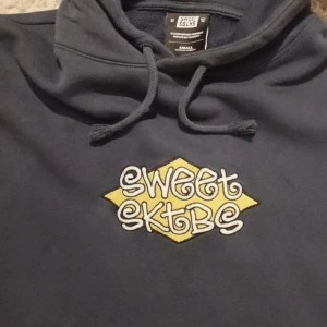 Mörkblå hoodie från Sweet SKTBS - Säljer en mörkblå hoodie från Sweet SKTBS med en cool gul och vit logga på bröstet. Hoodien har en klassisk känguruficka och justerbar huva med dragsko. Perfekt för en avslappnad stil!