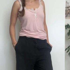 Rosa linne från Hunkemöller - Säljer ett supergulligt rosa linne från Hunkemöller. Det är gjort av mjukt ribbat material och har fina spetsdetaljer runt halsen och axelbanden. Linnet har också små knappar framtill som en söt detalj. Perfekt för sommaren eller som ett lager under en tröja!