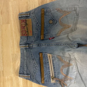 Blå jeans från Edwin - Säljer ett par snygga blå jeans från Edwin. De har en cool vintage look med slitningar och detaljer i brunt. Jeansen har en dragkedja på bakfickan och en knapp på den andra bakfickan. Perfekta för en avslappnad och stilren look!