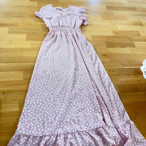 Rosa långklänning med vita prickar - Säljer en superfin rosa långklänning med vita prickar. Klänningen har korta ärmar med volangdetaljer och en smockad midja som ger en snygg passform. 