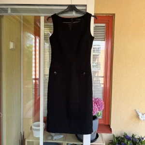 Svart klänning från H&M - Säljer en svart klänning från H&M i storlek 34. Liten i storlek. Klänningen är ärmlös och har en v-ringad hals. Den har en figursydd passform och två dekorativa fejkfickor med knappar framtill. Perfekt för kontoret.
