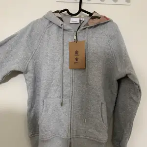 Säljer en snygg och bekväm grå hoodie från Burberry. Den har en dragkedja framtill, två fickor och en justerbar huva med snören. Perfekt för vardagsbruk eller som ett extra lager under kallare dagar. Hoodien är i nyskick och har aldrig använts.