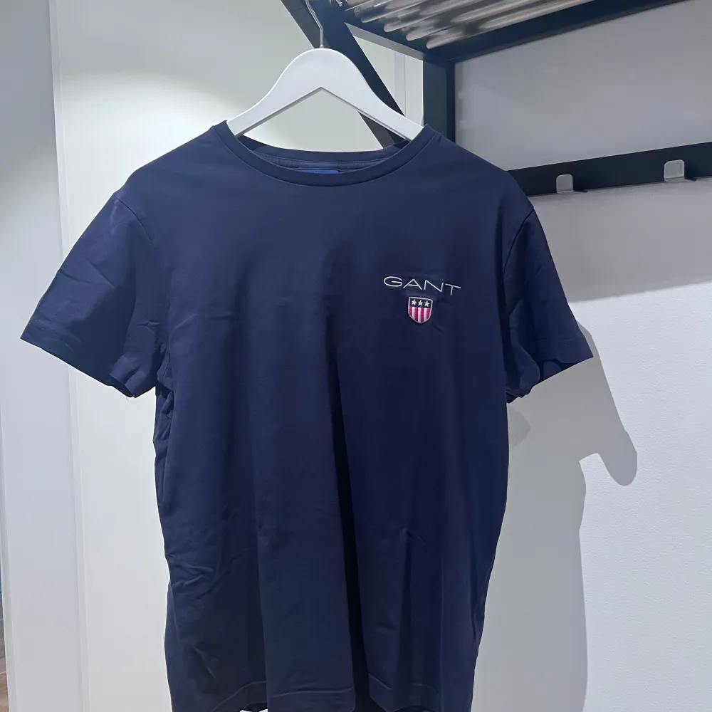 Säljer en snygg mörkblå t-shirt från GANT. Den har en liten logga på bröstet med texten 'GANT' och en sköld. Perfekt för en casual look eller för att bara chilla i. T-shirten är i bra skick och känns riktigt skön att ha på sig.. T-shirts.