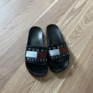 Säljer ett par svarta sandaler från Tommy Hilfiger. De har en bred rem över foten med Tommy Hilfiger-loggan i vitt, rött och blått. Sandalerna är i syntetmaterial och är i bra skick. Perfekta för sommaren!