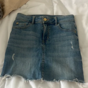 Jeanskjol med slitningar - Säljer en snygg jeanskjol i blå denim med slitningar. Kjolen har en rå kant nertill och klassisk design. 