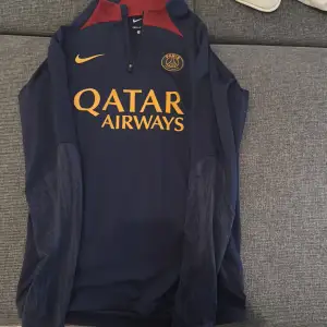 Säljer en långärmad Paris Saint-Germain fotbollströja från Nike. Tröjan är mörkblå med röda detaljer och har en gul Nike-logga samt Qatar Airways som sponsor. Den är i slim fit och tillverkad med Dri-FIT material för att hålla dig torr och bekväm. Perfekt för både träning och vardagsbruk!