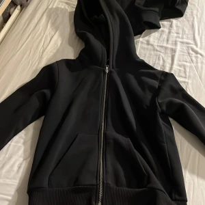 Svart hoodie med dragkedja - Säljer en stilren svart hoodie med dragkedja och huva. Den har långa ärmar och ribbade muddar vid ärmslut och nederkant. Perfekt för en avslappnad look.