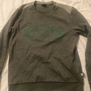 Grå sweatshirt från Hugo Boss - Snygg grå sweatshirt från Hugo Boss med grönt BOSS-tryck på framsidan. Tröjan har långa ärmar och en klassisk rund halsringning. Perfekt för en stilren och avslappnad look. Aldrig använt skick 10/10