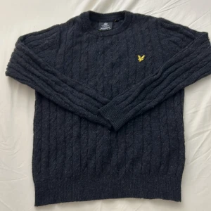 Mörkgrå tröja från Lyle & Scott - Säljer en stilren mörkgrå tröja från Lyle & Scott med kabelstickat mönster. Tröjan har en rund halsringning och långa ärmar. Den ikoniska loggan i gult är broderad på bröstet. Perfekt för kyliga dagar!
