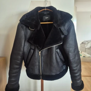 Svart bikerjacka med pälsdetaljer - Snygg svart skinnjacka från Bikbok med pälsdetaljer på kragen och ärmslut. Jackan har en asymmetrisk dragkedja och bälte i midjan för en cool look. Perfekt för kyliga dagar när du vill se extra stilren ut. Använd några tillfällen endast så fint skick!