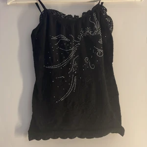 Svart topp med spets och pärlor - Snygg svart topp med spetsdetaljer och pärlor som bildar ett elegant mönster. Perfekt för en kväll ute eller en speciell tillställning. Toppen har en tight passform och är tillverkad i ett mjukt material. en detalj har trillat av i mittan av tröjan men jag tyckte den va finare utan! annars är den i nyskick