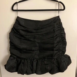 Svart kjol med volangdetaljer - Snygg svart kjol med volangdetaljer och rynkad design. Kjolen har en kort längd och är perfekt för att ge en elegant touch till din outfit. Den har en subtil glans i materialet som ger en lyxig känsla.