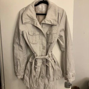 Beige jacka från H&M L.O.G.G. - Säljer en stilren beige jacka från H&M L.O.G.G. med knappar och bälte i midjan. Jackan har långa ärmar och flera praktiska fickor framtill. Perfekt för vår och höst!