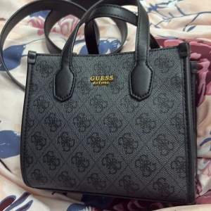 Svart handväska från Guessny - Snygg svart handväska från Guess med ett diskret mönster och guldfärgade detaljer. Väskan har dubbla handtag och en avtagbar axelrem för extra flexibilitet. Perfekt för att bära dina nödvändigheter med stil.