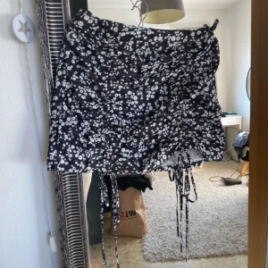 Svart blommig kjol med snörning - Säljer en snygg svart kjol med vita blommor och snörningsdetaljer framtill. Kjolen har en kort längd och är perfekt för en trendig look. Den har en åtsittande passform. 
