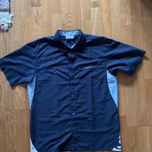 Mörkblå skjorta från Quiksilver - Säljer en mörkblå skjorta från Quiksilver med korta ärmar och knappar framtill. Skjortan har en bröstficka med Quiksilver-loggan broderad. Perfekt för en avslappnad stil.