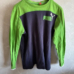 Grön och svart långärmad tröja från Puma - Säljer en cool långärmad tröja från Puma i grönt och svart. Tröjan har en klassisk rund hals och Puma-loggan tryckt på bröstet. Perfekt för en avslappnad stil.