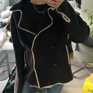 Vårjacka - Snygg svart kappa med vita kontrastsömmar och dubbelknäppning. Jackan har en klassisk krage och långa ärmar. Perfekt för en stilren look.