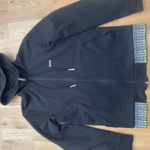 Svart huvtröja från Boss - Säljer en stilren svart huvtröja från Boss med dragkedja och huva. Tröjan har långa ärmar och ribbade muddar. Detaljer i form av Boss-logga på bröstet och mönstrad kant nedtill