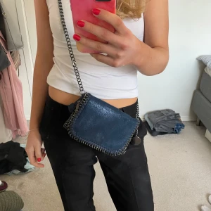 Blå axelväska med kedjedetaljer - Snygg blå axelväska med kedjedetaljer och dragkedja. Väskan har en unik design med en kedja som axelrem och dekorativa sömmar längs kanterna. Perfekt för att ge din outfit en cool touch.