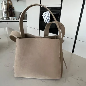 Beige axelväska i mocka - Snygg beige axelväska i mocka med stilren design. Väskan har en praktisk storlek och är perfekt för att bära med sig det viktigaste. Den har både handtag och en axelrem för flexibel användning.