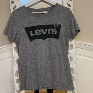 Grå t-shirt från Levi's - Säljer en grå t-shirt från Levi's med deras ikoniska logga i svart på bröstet. T-shirten är kortärmad och tillverkad i ett mjukt bomullsmaterial. Perfekt för en avslappnad stil.