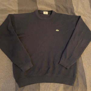Mörkblå stickad tröja från Lacoste - Säljer en mörkblå stickad tröja från Lacoste med det klassiska krokodilmärket på bröstet. Tröjan har långa ärmar och ribbstickade muddar vid ärmslut och nederkant. Perfekt för kyligare dagar! Lite lösa trådar i ärmsluten och minimalt med fluff saker men annars perfekt skick