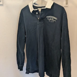 Mörkblå pikétröja från Peak Performance - Säljer en stilren mörkblå pikétröja från Peak Performance med vit krage och knappdetaljer. Tröjan har långa ärmar och ett tryck på bröstet. Perfekt för en avslappnad och sportig look.