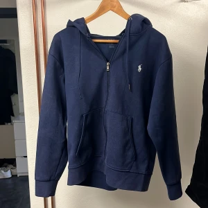 Mörkblå hoodie från Ralph Lauren - Säljer en snygg mörkblå hoodie från Ralph Lauren med dragkedja och det klassiska broderade logotypen på bröstet. Tröjan har en bekväm passform och är perfekt för kyliga dagar. Den har en praktisk huva och fickor framtill.
