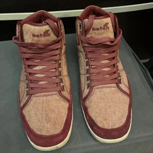 Rödmelerade sneakers från Boxfresh - Snygga rödmelerade sneakers från Boxfresh med hög design och snörning. Skorna har en kombination av textil och mocka, vilket ger dem en unik look. Perfekta för en avslappnad stil.