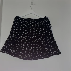 Svart blommig kjol - Svart kjol med små vita blommor. Har ett snöre som går runt och knyts på sidan. Finns dragkedja och innershorts i samma mönster. 💐