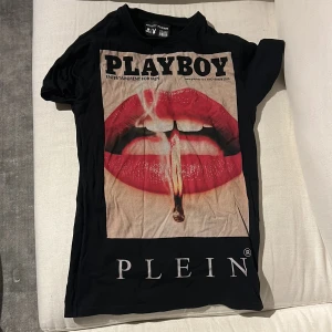 Svart t-shirt med tryck från Philipp Plein - Cool svart t-shirt från Philipp Plein med ett stort tryck av röda läppar och texten 'PLAYBOY' på framsidan. T-shirten är kortärmad och har en modern design. Perfekt för dig som vill sticka ut med stil.
