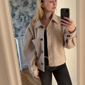 Beige jacka från NA-KD - Säljer en snygg beige jacka från NA-KD med knappar framtill och stora fickor. Jackan har en oversized passform och långa ärmar. Har en svart fläck på höger arm