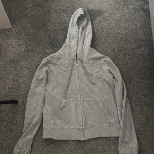 Grå hoodie från Juicy Couture - Säljer en stilren grå hoodie från Juicy Couture med dragkedja och huva. Tröjan har långa ärmar och en liten logotyp på bröstet. Perfekt för en avslappnad look.