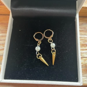 Pearl spike - Snygga guldörhängen med vita pärlor och konformade hängen. Perfekta för att ge en edgy touch till din stil. De har en enkel låsmekanism som gör dem lätta att bära.