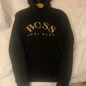 Svart hoodie från Hugo Boss - Snygg svart hoodie från Hugo Boss med stor logga i beige på framsidan. Tröjan har en klassisk design med huva och dragsko samt en praktisk ficka framtill. Perfekt för en stilren och bekväm look.