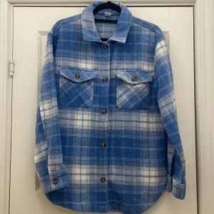 Blå och vit rutig overshirt - Säljer en snygg blå och vit rutig overshirt med långa ärmar och knappar framtill. Skjortan har två bröstfickor med knappar och är perfekt för en avslappnad stil. Materialet känns mjukt och bekvämt.