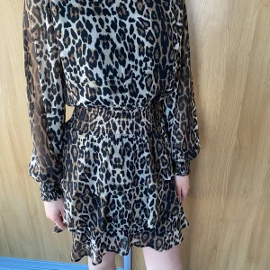 Leopardmönstrad klänning - Trendig leopardmönstrad klänning med långa ärmar och volangdetaljer. Perfekt för att sticka ut med stil. Den har en elastisk midja för en bekväm passform.