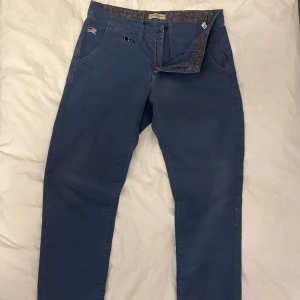 Blå chinos från Burberry - Snygga blå chinos från Burberry med broderad logga och stiliga detaljer. Storlek 46 Byxorna har en dragkedja och knappar samt mönstrade fickor. Perfekta för en stilren look. har en liten fläck på bakfickan. Syns inte lika mycket i verkligheten. 