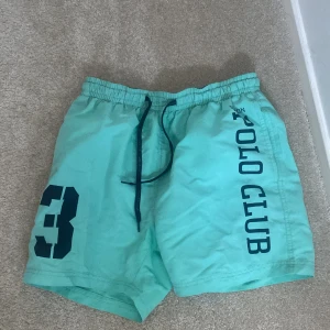 Turkosa shorts från Vinson Polo Club/badshorts - Snygga turkosa shorts från Vinson Polo Club med mörkblå snörning och tryck. Perfekta för en avslappnad stil med elastisk midja för extra komfort. Stort nummertryck på ena benet ger en sportig touch.