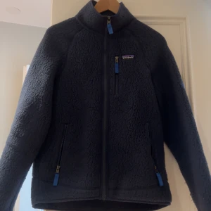 Mörkblå fleecejacka från Patagonia - Säljer en mörkblå fleecejacka från Patagonia med dragkedja framtill och två praktiska fickor med dragkedjor. Jackan har en hög krage och är perfekt för kyliga dagar. Den är både stilren och funktionell. 