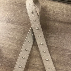 Beige bälte med pärldetaljer - Snyggt beige väskband från Don Donna med pärldetaljer. Tillverkat i skinn med en texturerad yta och silverfärgade spännen. Perfekt för att ge en elegant touch till din outfit.