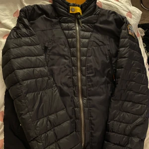 Svart quiltad jacka med dragkedja - Säljer en svart quiltad jacka från Parajumpers med dragkedja och hög krage. Jackan har långa ärmar och en gul detalj vid halsen. Perfekt för kyligare dagar.