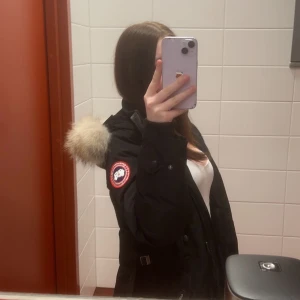 Svart parkajacka med pälsdetalj - Säljer en svart parkajacka från Canada Goose med en mysig pälsdetalj på huvan. Jackan har en dragkedja och knappar framtill samt en ikonisk logga på ärmen. Perfekt för kalla dagar!