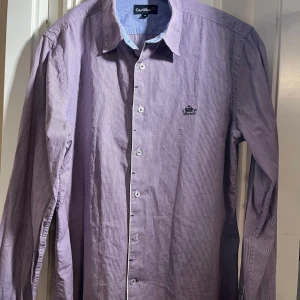 Lila randig skjorta från EastWest - Säljer en stilren lila skjorta från EastWest med diskret randigt mönster och broderad logga på bröstet. Skjortan har långa ärmar och knappar framtill. Perfekt för en snygg och avslappnad look.