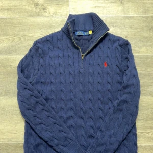 Mörkblå stickad tröja från Ralph Lauren - Säljer en snygg blå stickad tröja från Ralph Lauren med dragkedja vid halsen. Tröjan har ett klassiskt flätmönster och en röd broderad logotyp på bröstet. Storleken är XL men skulle säga att det passar till L. Jag själv har storlek L 