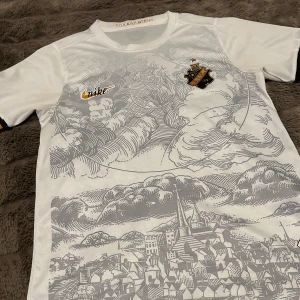 Vit AIK fotbollströja från Nike - Snygg vit AIK fotbollströja från Nike med ett unikt mönster av en stadssilhuett i grått. Tröjan har korta ärmar och AIK:s emblem på bröstet. Perfekt för alla AIK-fans som vill visa sitt stöd med stil.